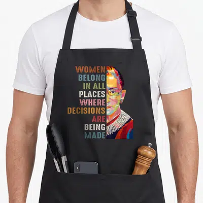 Belong novelty apron - Stacked multicolored text next to a stylized portrait of a woman wearing glasses and a lace — shown on a black cotton canvas apron against a white background