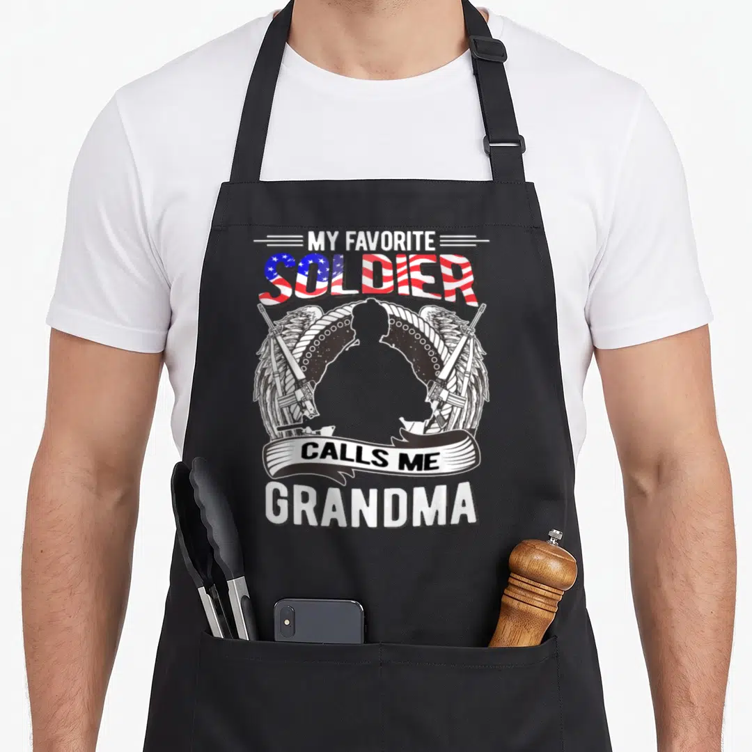 Army grandma apron - My Favorite Soldier Calls Me Grandma text with flag letters and soldier silhouette flanked by two — shown on a black cotton canvas apron against a white background