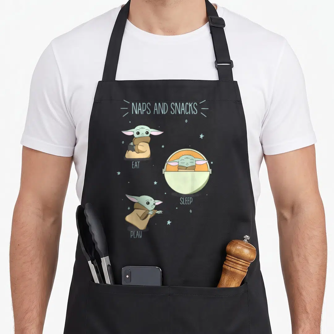 Snack nap apron - Grogu cartoon eating sleeping playing with NAPS AND SNACKS text and stars — shown on a black cotton canvas apron against a white background