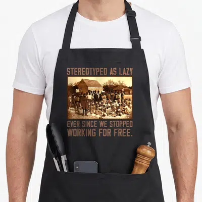 Stereotyped a's aprons - Sepia group photo with orange text stereotyped as lazy ever since we stopped working for free — shown on a black cotton canvas apron against a white background