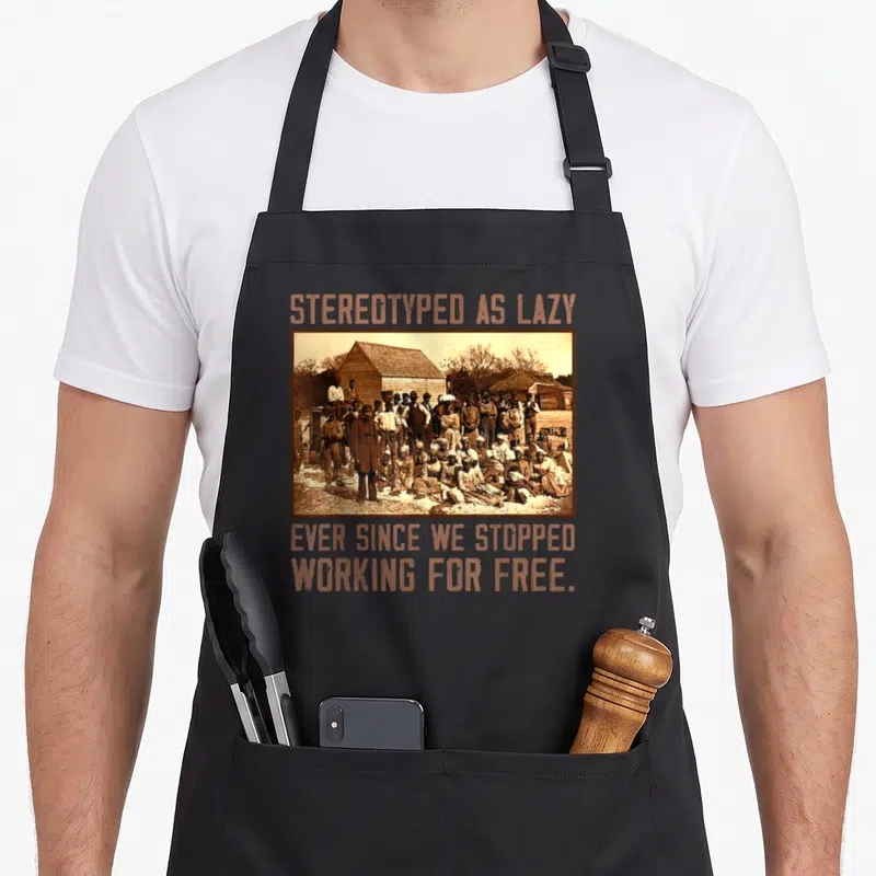 Stereotyped a's aprons - Sepia group photo with orange text stereotyped as lazy ever since we stopped working for free — shown on a black cotton canvas apron against a white background