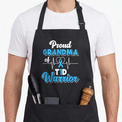 T1d grandma apron - Stacked blue and white text reading Proud GRANDMA of T1D Warrior with a heartbeat ribbon graphic — shown on a black cotton canvas apron against a white background