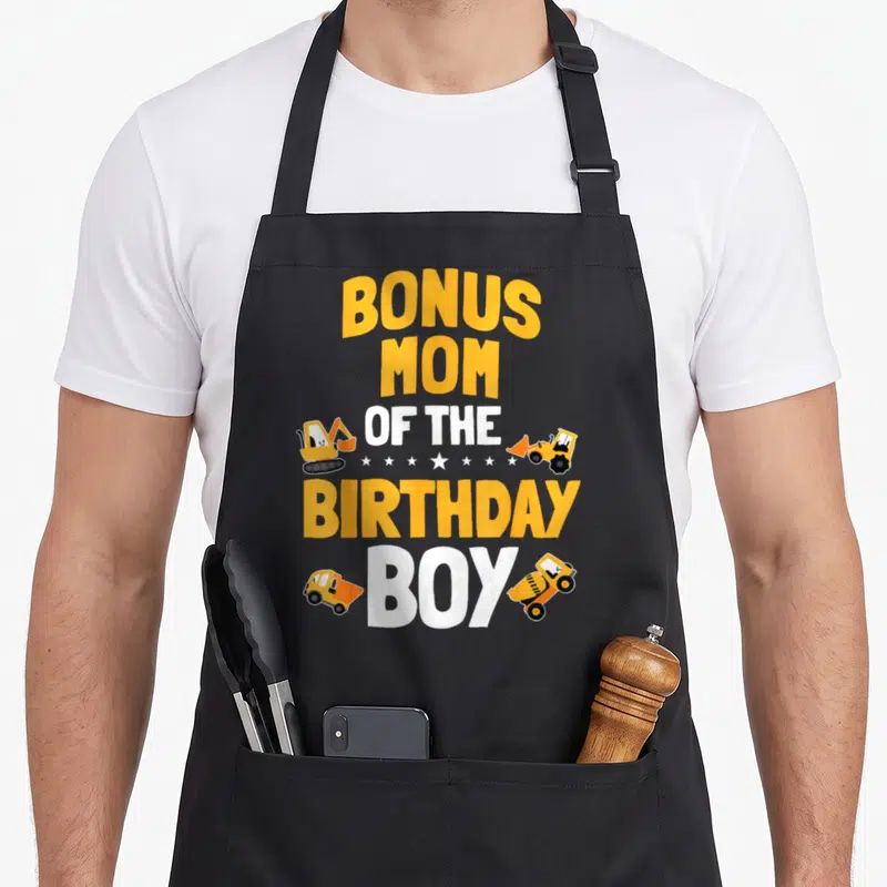 Bonus construction worker apron - Yellow and white Bonus Mom of the Birthday Boy text with excavator, bulldozer, dump — shown on a black cotton canvas apron against a white background