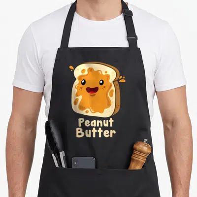 Halloween butter apron - Smiling cartoon toast with orange spread and text reading Peanut Butter — shown on a black cotton canvas apron against a white background
