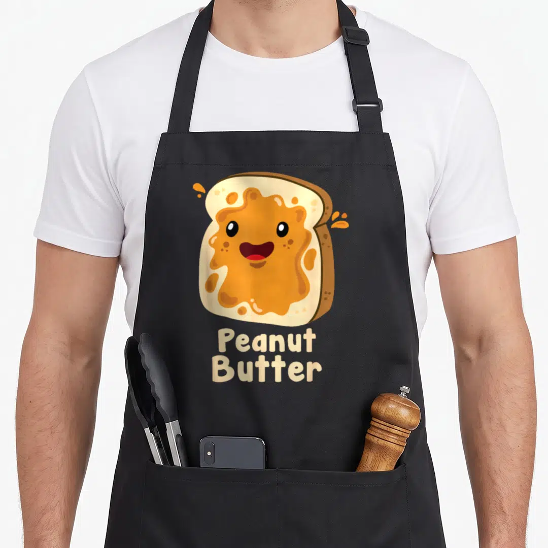 Halloween butter apron - Smiling cartoon toast with orange spread and text reading Peanut Butter — shown on a black cotton canvas apron against a white background