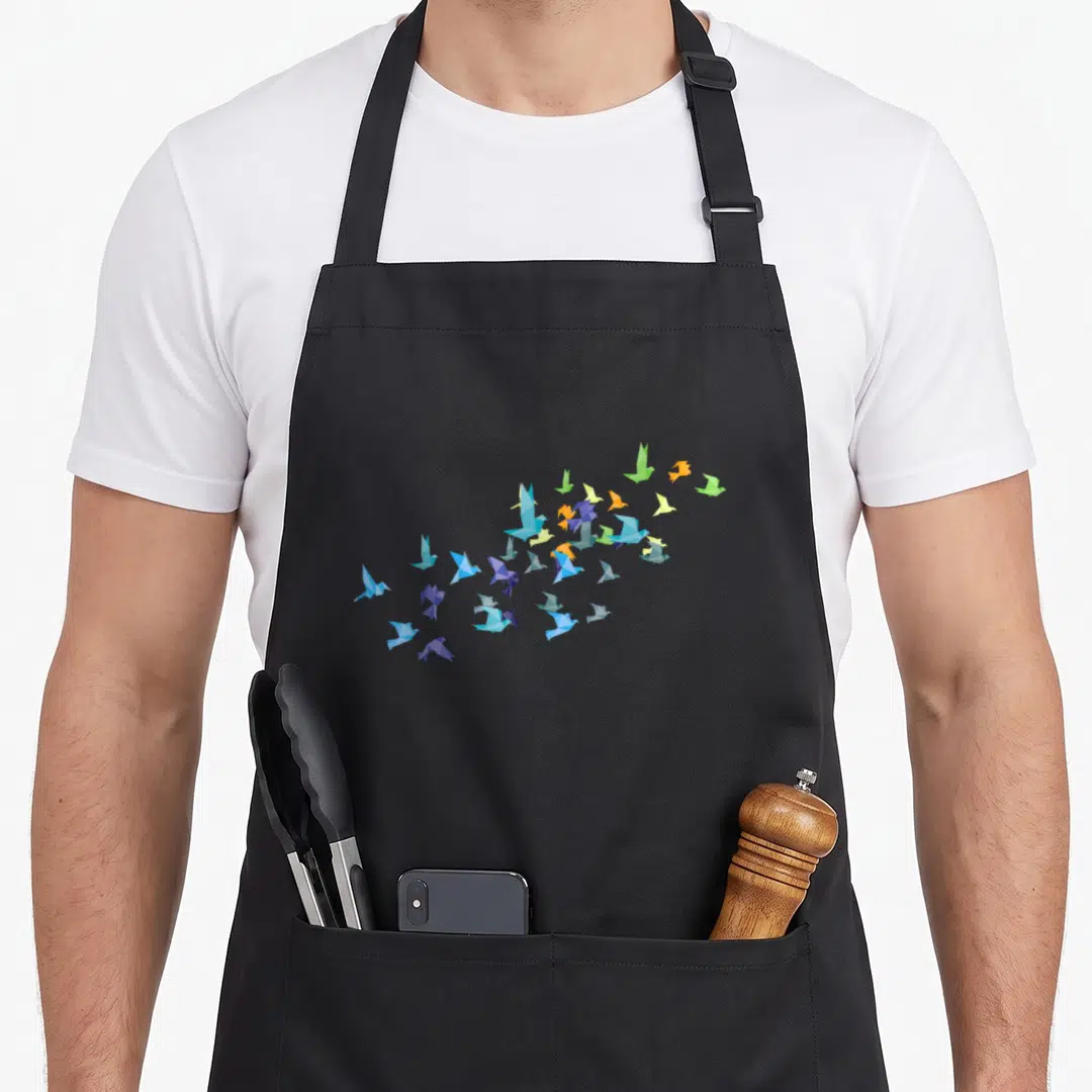 Crane paper apron - Geometric origami birds in blue, purple, yellow, orange, and green flying in a cluster formation — shown on a black cotton canvas apron against a white background