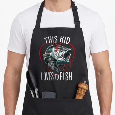 Funny fishing fish apron - Cartoon fish inside red heart outline with white text reading THIS KID LOVES TO FISH — shown on a black cotton canvas apron against a white background