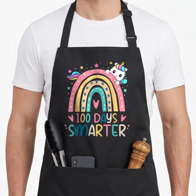 100th unicorn apron - Rainbow with peeking unicorn and 100 DAYS SMARTER text in pink yellow blue white — shown on a black cotton canvas apron against a white background