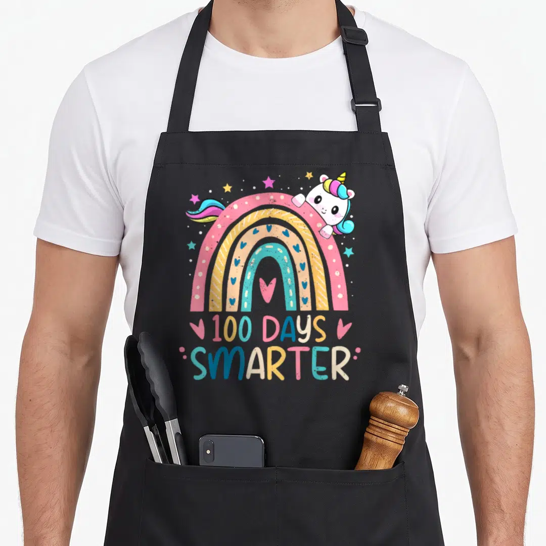 100th unicorn apron - Rainbow with peeking unicorn and 100 DAYS SMARTER text in pink yellow blue white — shown on a black cotton canvas apron against a white background