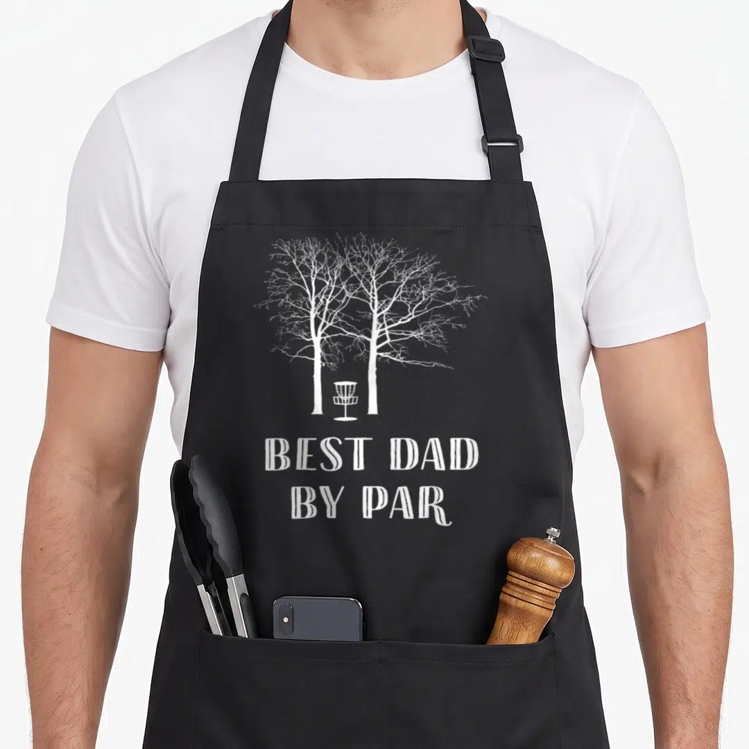 Disc par golf apron - White disc golf basket between two trees with text best dad by par — shown on a black cotton canvas apron against a white background