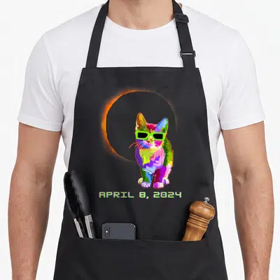 Solar cat wearing apron - Multicolored cartoon cat with neon green sunglasses in front of orange eclipse ring and date — shown on a black cotton canvas apron against a white background
