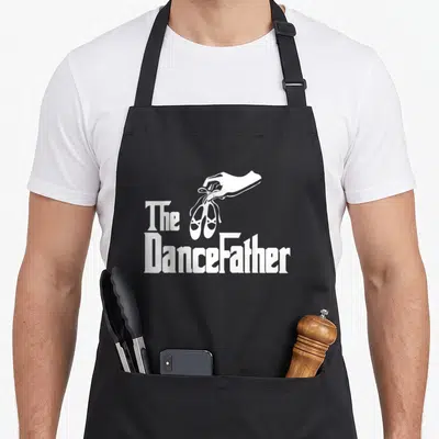 Dancefather dad performance apron - White hand holding ballet shoes with The DanceFather text on black fabric — shown on a black cotton canvas apron against a white background