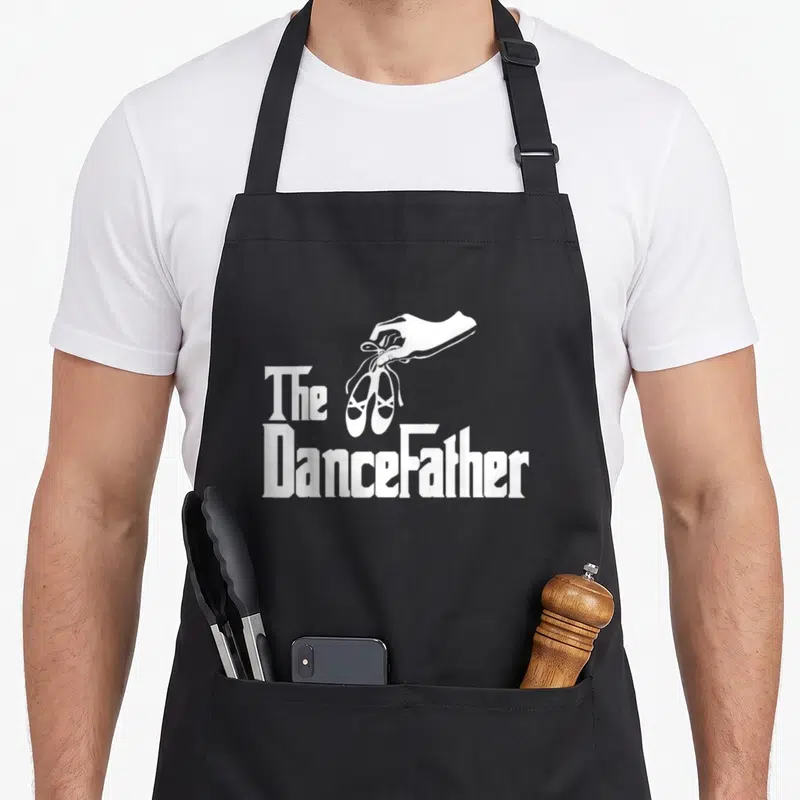 Dancefather dad performance apron - White hand holding ballet shoes with The DanceFather text on black fabric — shown on a black cotton canvas apron against a white background