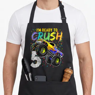 5th birthday truck apron - Monster truck jumping over white number 5 with I'm ready to crush text and confetti — shown on a black cotton canvas apron against a white background