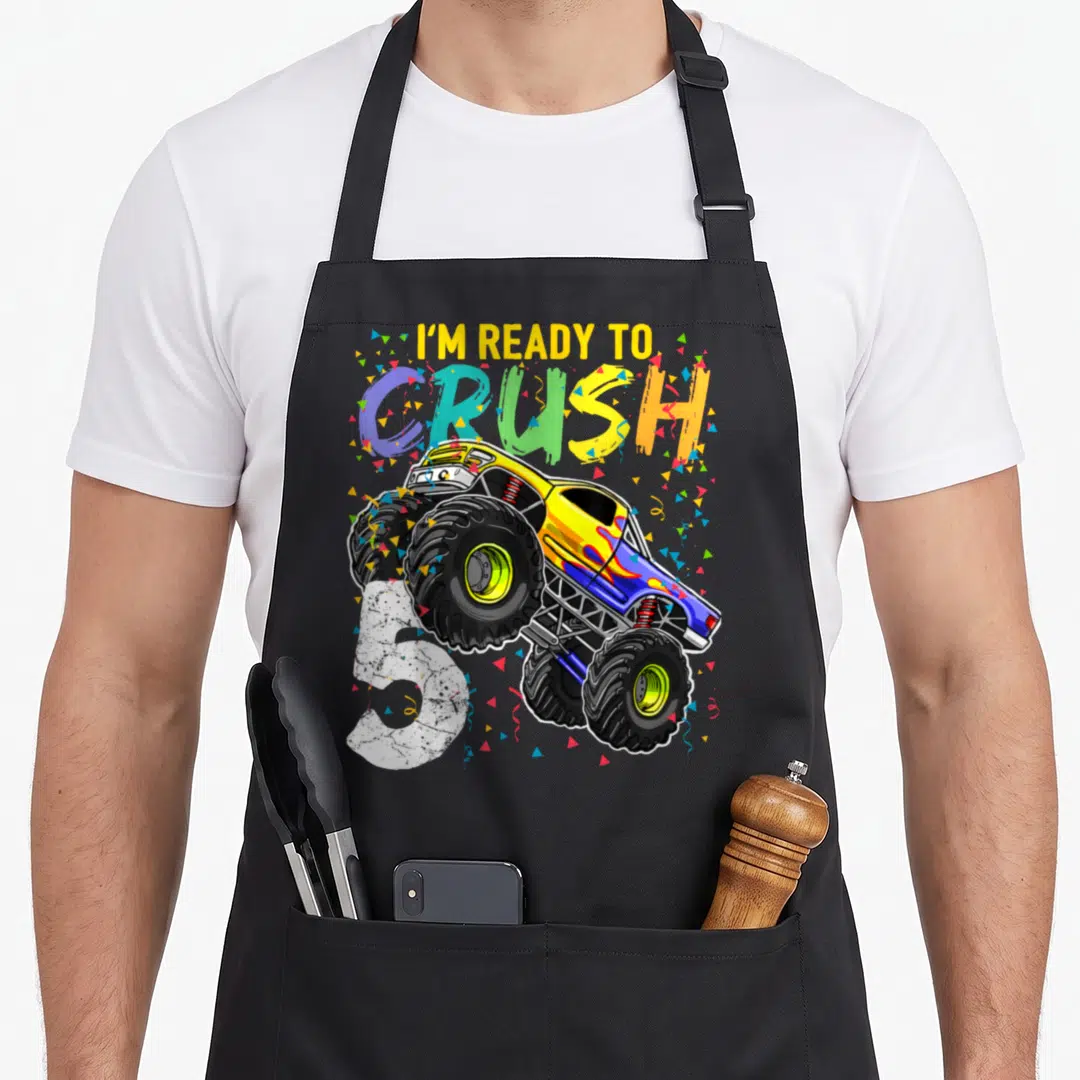 5th birthday truck apron - Monster truck jumping over white number 5 with I'm ready to crush text and confetti — shown on a black cotton canvas apron against a white background