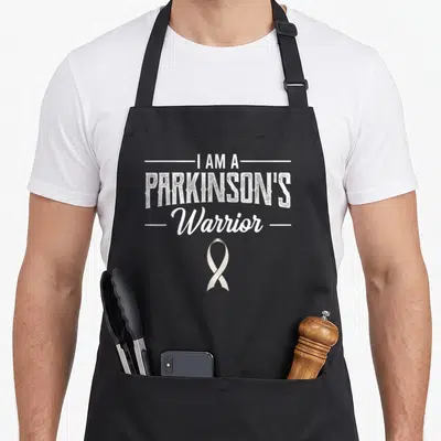 Idiopathic ribbon apron - White stacked text I Am A Parkinson's Warrior above a centered ribbon on black fabric — shown on a black cotton canvas apron against a white background