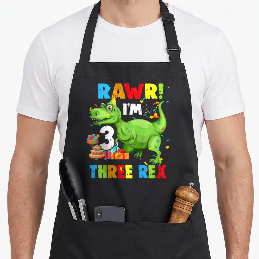 Three chef outfit apron - Green T-Rex party hat cake balloons THREE REX text — shown on a black cotton canvas apron against a white background