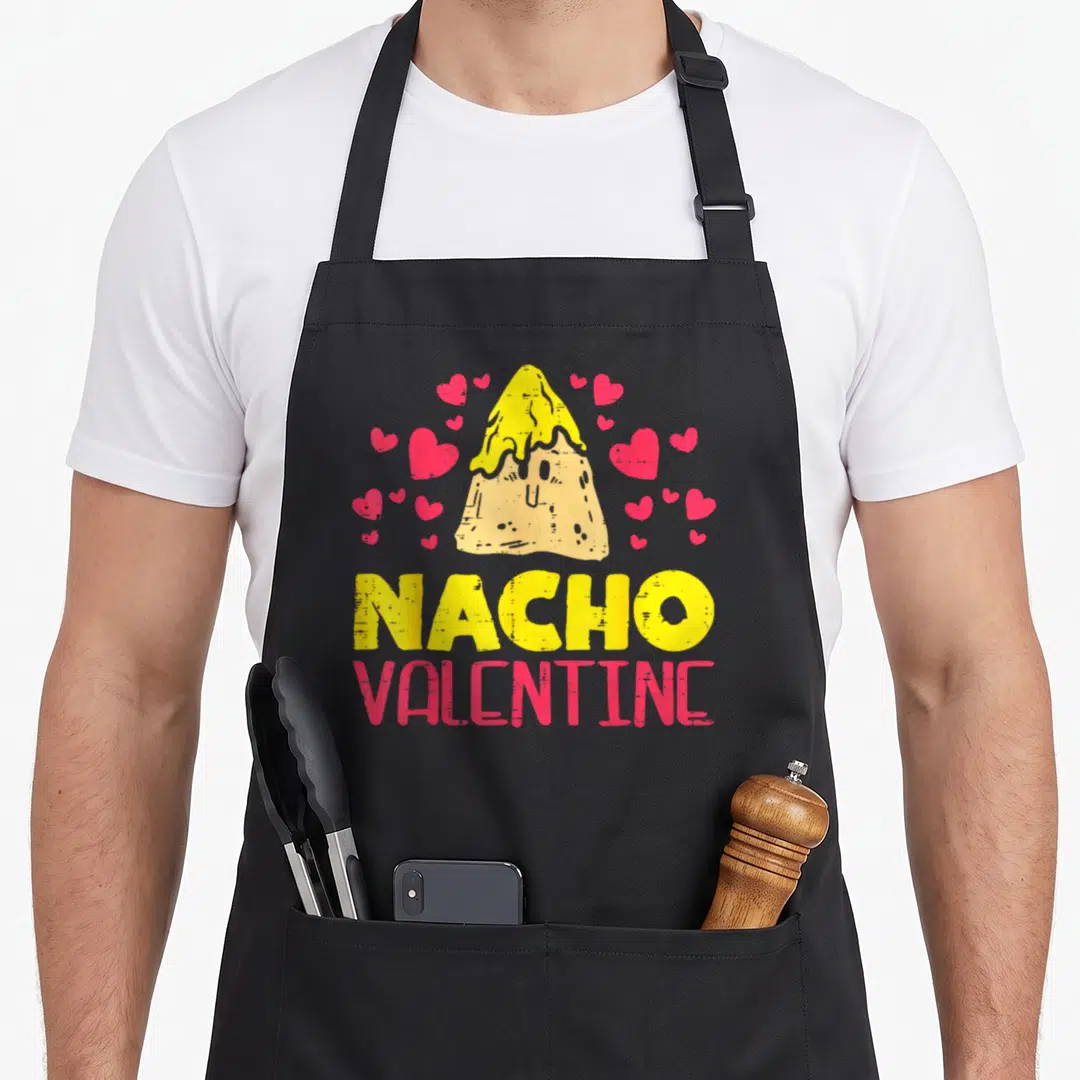 Nacho valentine's day apron - Cartoon nacho chip with yellow cheese and pink hearts above NACHO VALENTINE text — shown on a black cotton canvas apron against a white background