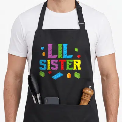Lil block & apron - LIL SISTER text formed by colorful toy bricks with scattered pieces — shown on a black cotton canvas apron against a white background