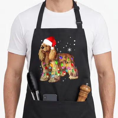 Christmas cocker spaniel apron - Golden brown cocker spaniel in red santa hat wrapped in multicolor lights — shown on a black cotton canvas apron against a white background