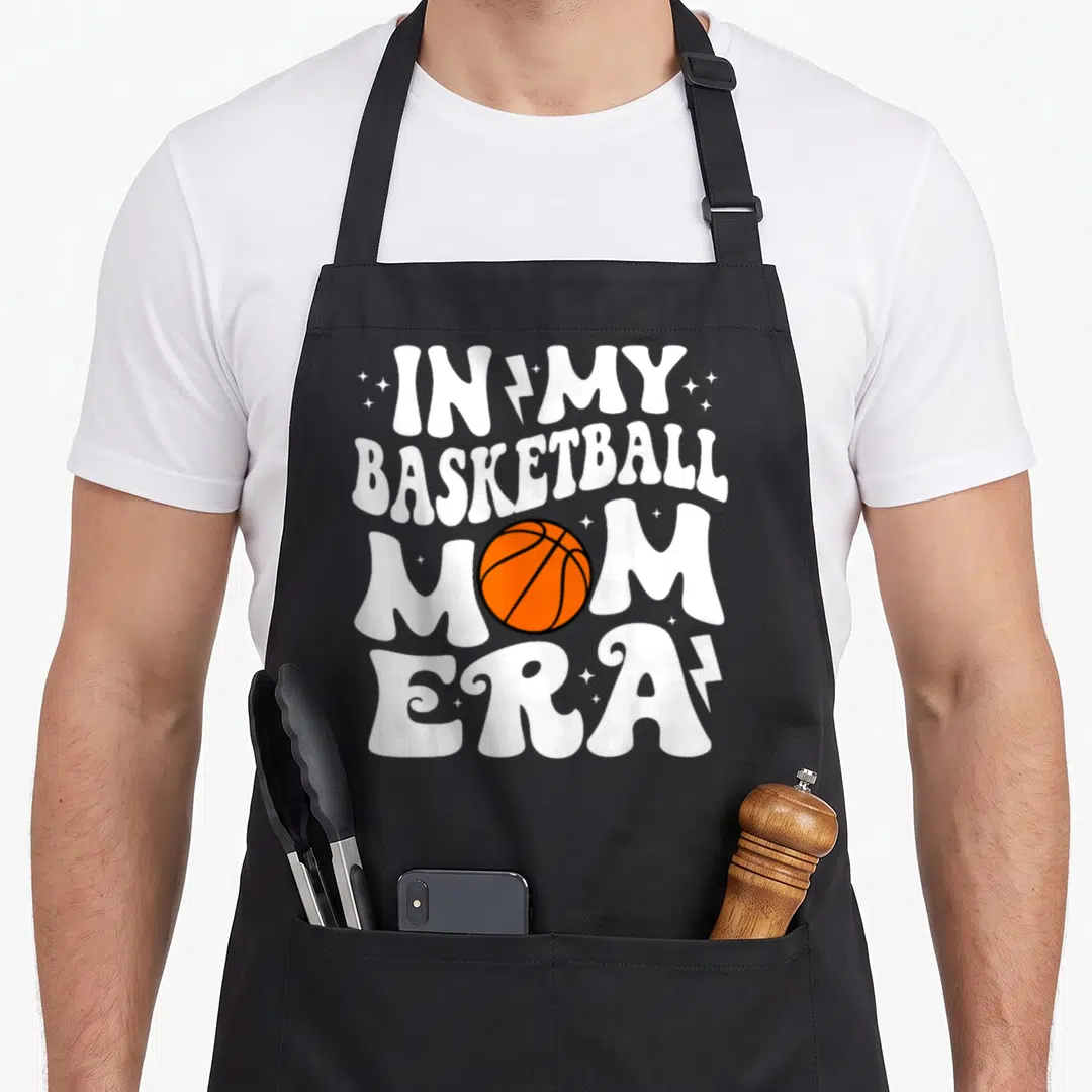 Era basketball mom apron - White stacked text reading IN MY BASKETBALL MOM ERA with orange basketball and star graphics — shown on a black cotton canvas apron against a white background