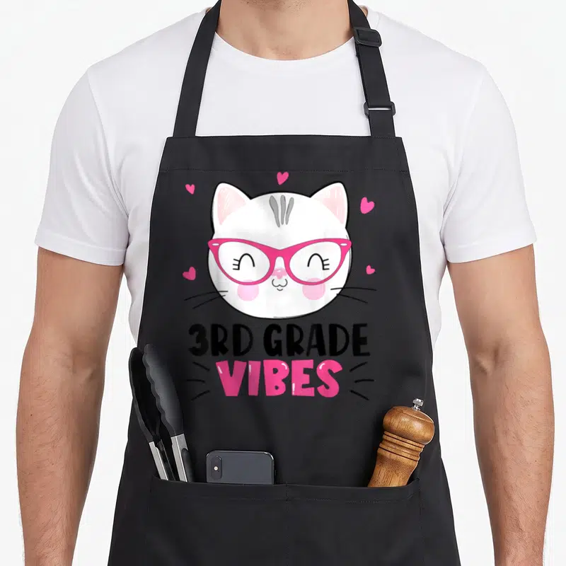 3rd cat apron - Cartoon white cat with pink glasses and hearts above 3RD GRADE VIBES text — shown on a black cotton canvas apron against a white background