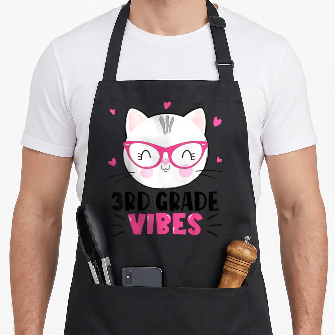 3rd cat apron - Cartoon white cat with pink glasses and hearts above 3RD GRADE VIBES text — shown on a black cotton canvas apron against a white background