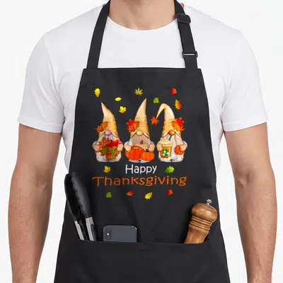 Gnome thanksgiving aprons - Three cartoon gnomes holding leaves pumpkins and a cup surrounded by scattered autumn — shown on a black cotton canvas apron against a white background