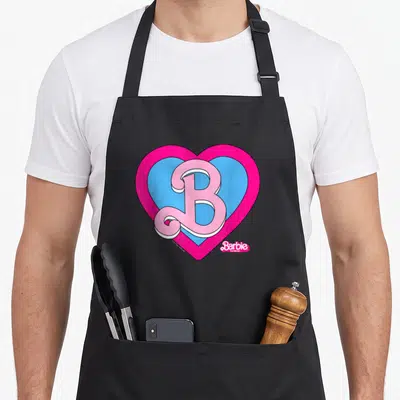 Movie barbiecore apron - Pink cursive B inside blue and pink hearts with white outlines and movie title text — shown on a black cotton canvas apron against a white background
