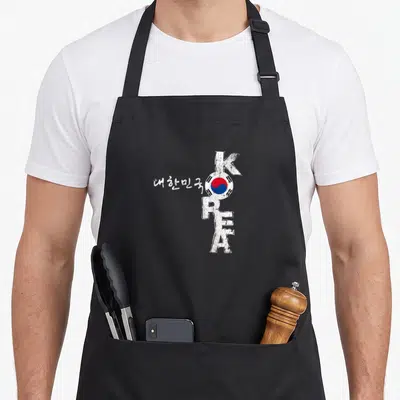 Korea korean apron - Vertical KOREA text with circular flag replacing O and white Korean characters to the left — shown on a black cotton canvas apron against a white background