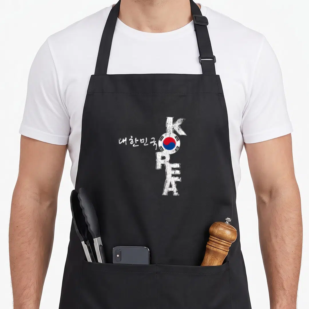 Korea korean apron - Vertical KOREA text with circular flag replacing O and white Korean characters to the left — shown on a black cotton canvas apron against a white background