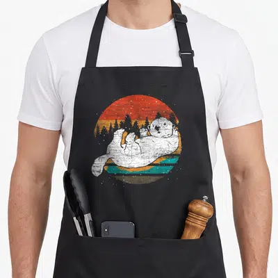 Vintage sea otter apron - White otter floating in front of red orange teal striped circle with pine forest silhouette — shown on a black cotton canvas apron against a white background