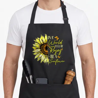 Rose sunflower apron - Yellow watercolor sunflower next to stacked text with butterflies and stars — shown on a black cotton canvas apron against a white background