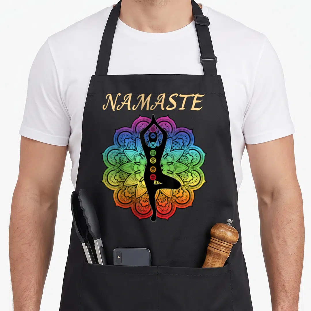 Chakra yoga apron - Namaste text above tree pose silhouette inside rainbow mandala with vertical chakra symbols — shown on a black cotton canvas apron against a white background