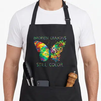 Broken psychiatrist apron - Watercolor butterfly with floral wing and text BROKEN CRAYONS STILL COLOR — shown on a black cotton canvas apron against a white background