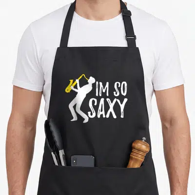 Jazz saxophone lover apron - White saxophonist silhouette playing yellow instrument beside stacked text IM SO SAXY — shown on a black cotton canvas apron against a white background