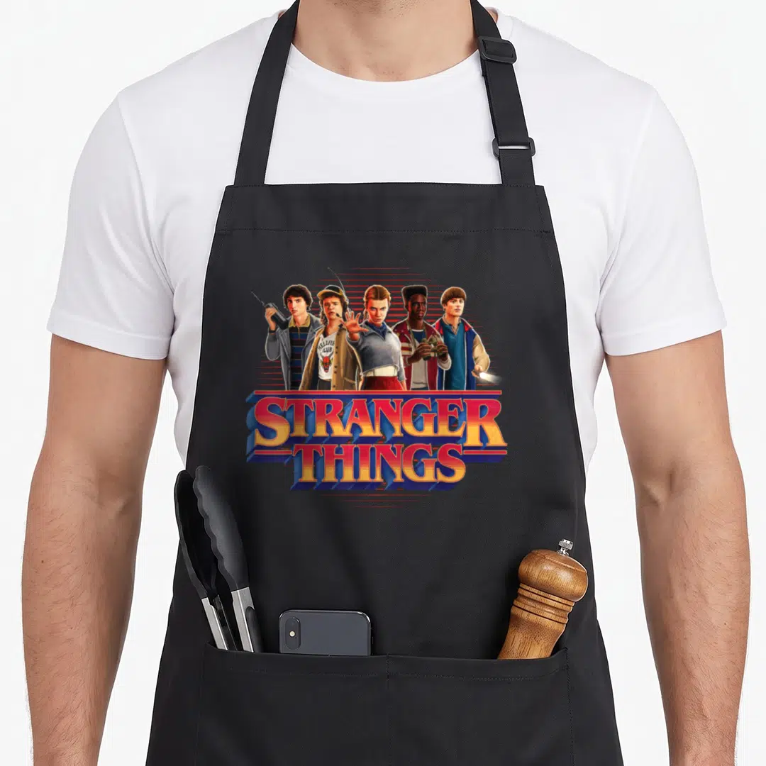 Season 80s apron - Five characters standing above stacked STRANGER THINGS text in red, blue, and yellow retro style — shown on a black cotton canvas apron against a white background