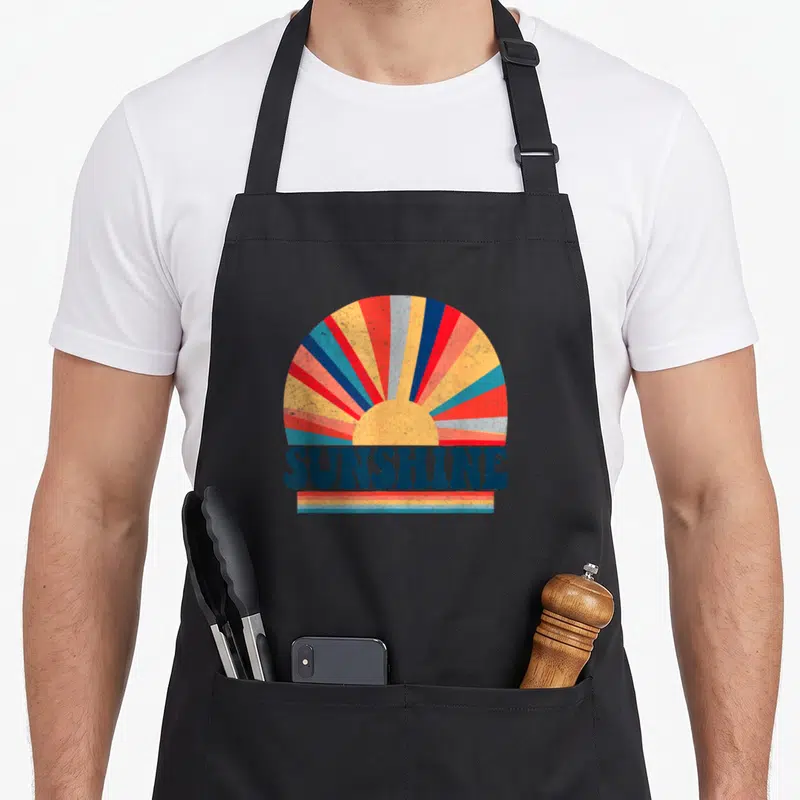Hippie 70s apron - Retro sunburst with SUNSHINE text and three colored stripes below — shown on a black cotton canvas apron against a white background