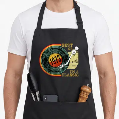 48th vinyl apron - Retro vinyl record with tonearm and 1973 text on striped background — shown on a black cotton canvas apron against a white background