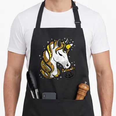 Candy corn halloween apron - Cartoon unicorn with candy corn mane holding a pumpkin candy bucket — shown on a black cotton canvas apron against a white background