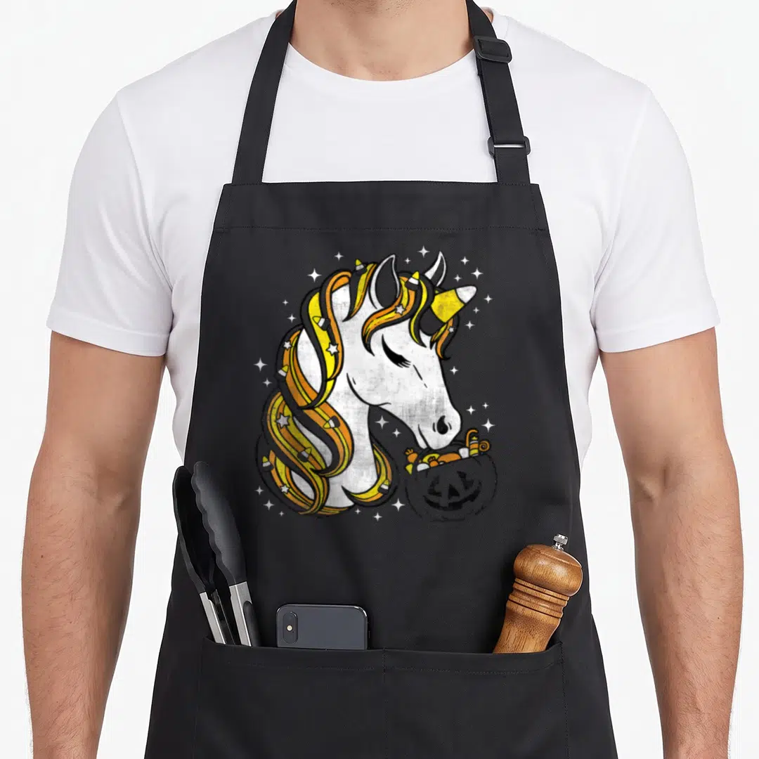 Candy corn halloween apron - Cartoon unicorn with candy corn mane holding a pumpkin candy bucket — shown on a black cotton canvas apron against a white background