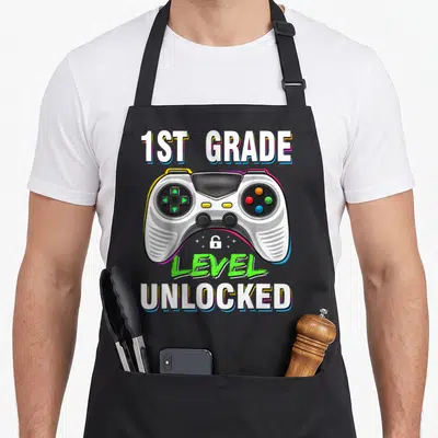 1st level apron - Game controller with 1st Grade Level Unlocked text and small lock icon — shown on a black cotton canvas apron against a white background
