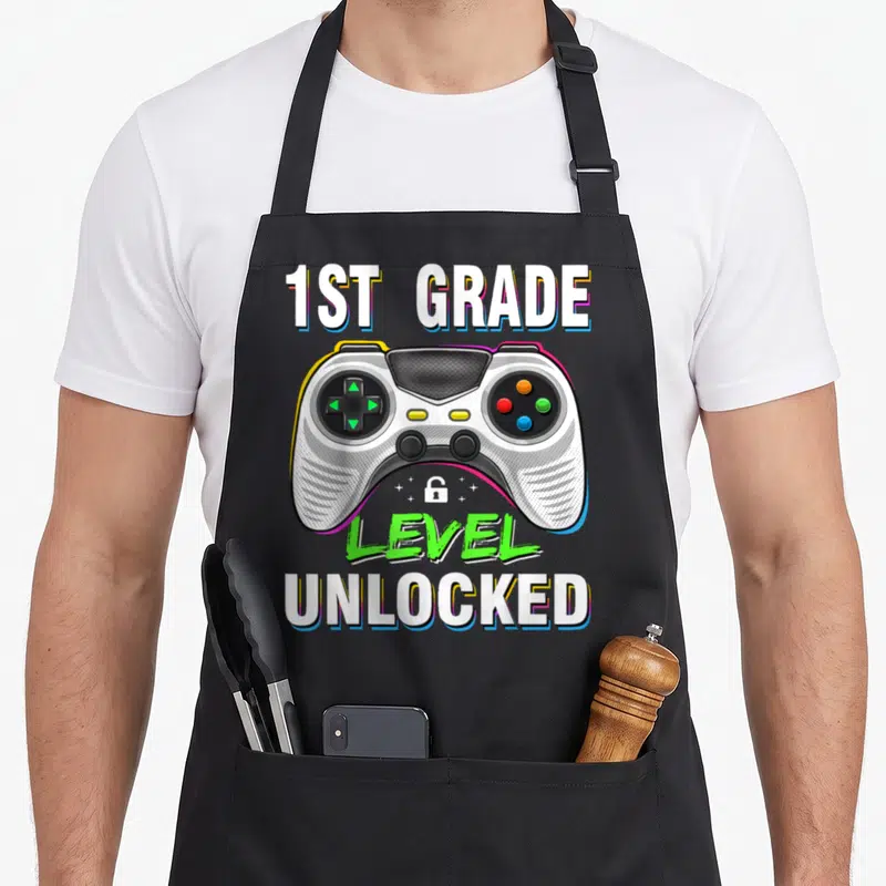 1st level apron - Game controller with 1st Grade Level Unlocked text and small lock icon — shown on a black cotton canvas apron against a white background