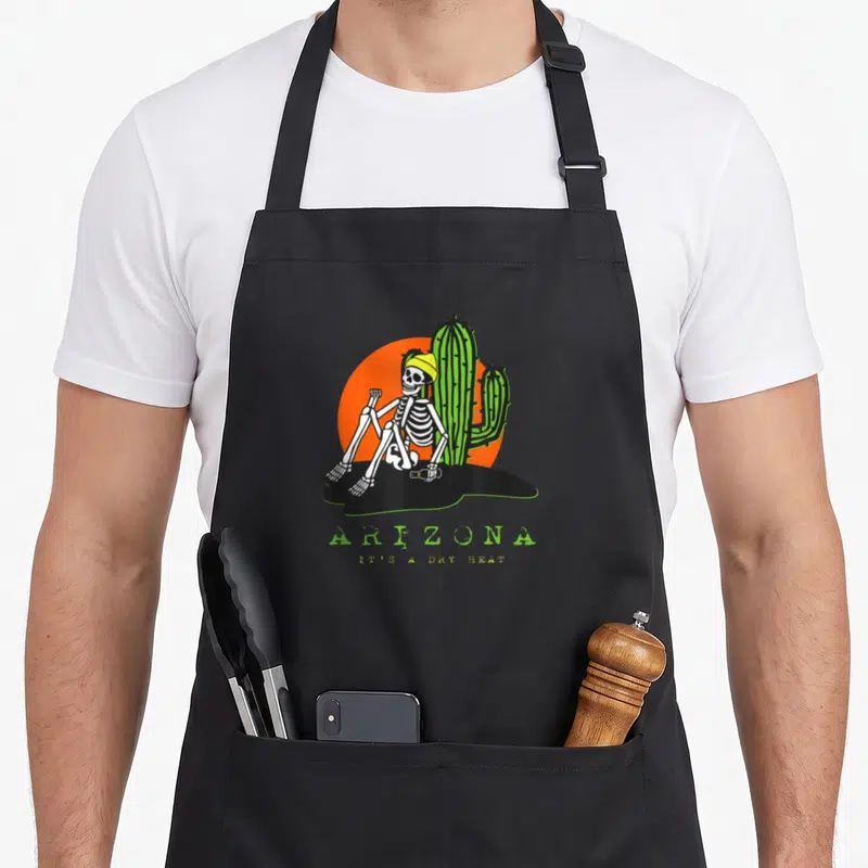Arizona skeleton apron - Cartoon skeleton in beanie sitting by cactus with ARIZONA IT'S A DRY HEAT text — shown on a black cotton canvas apron against a white background