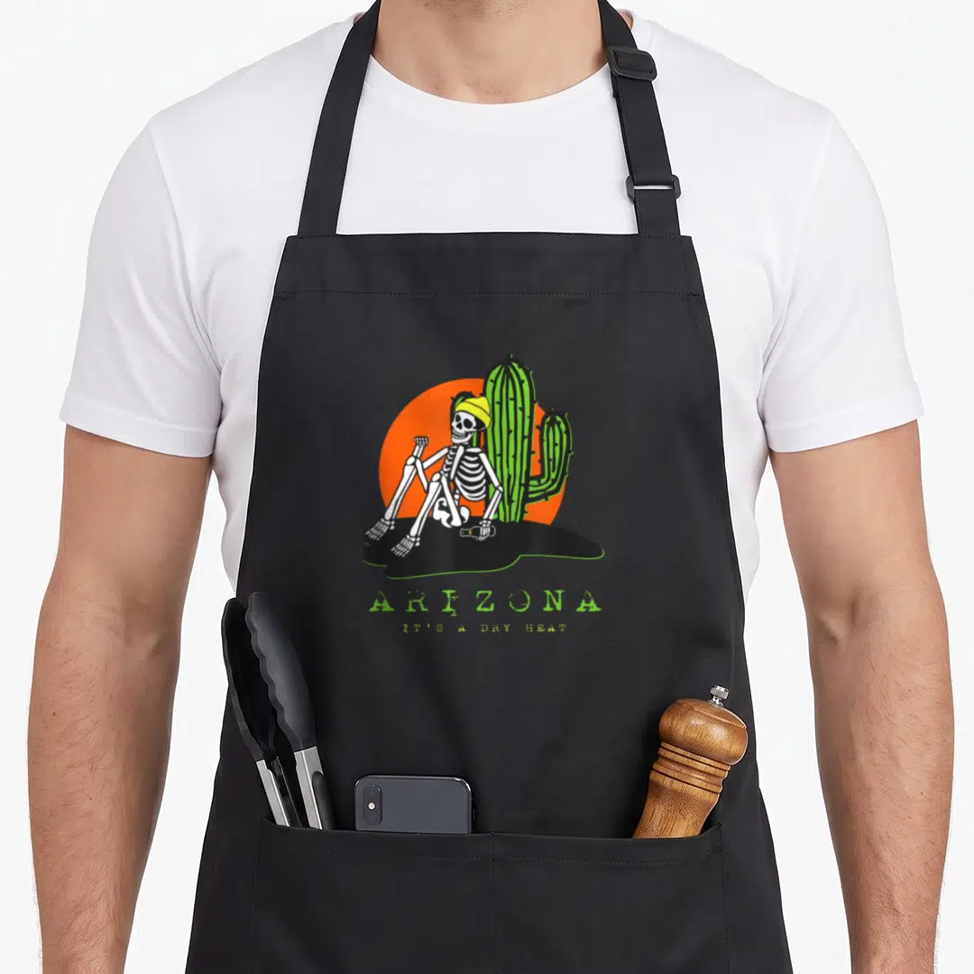 Arizona skeleton apron - Cartoon skeleton in beanie sitting by cactus with ARIZONA IT'S A DRY HEAT text — shown on a black cotton canvas apron against a white background