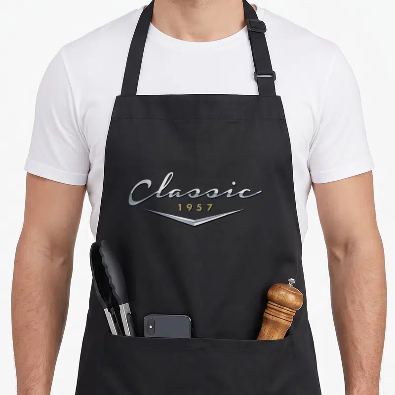Birthday husband apron - Classic 1957 text in silver script and gold numbers with curved underline on black fabric — shown on a black cotton canvas apron against a white background