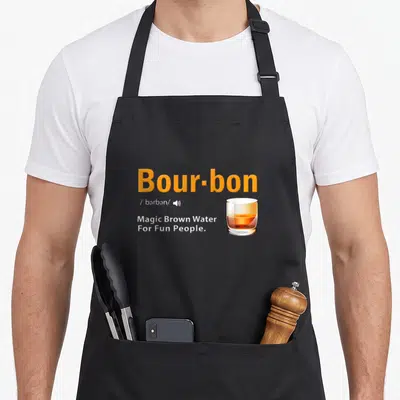 Bourbon magic apron - Orange text Bour-bon with glass of amber liquid and white definition text — shown on a black cotton canvas apron against a white background