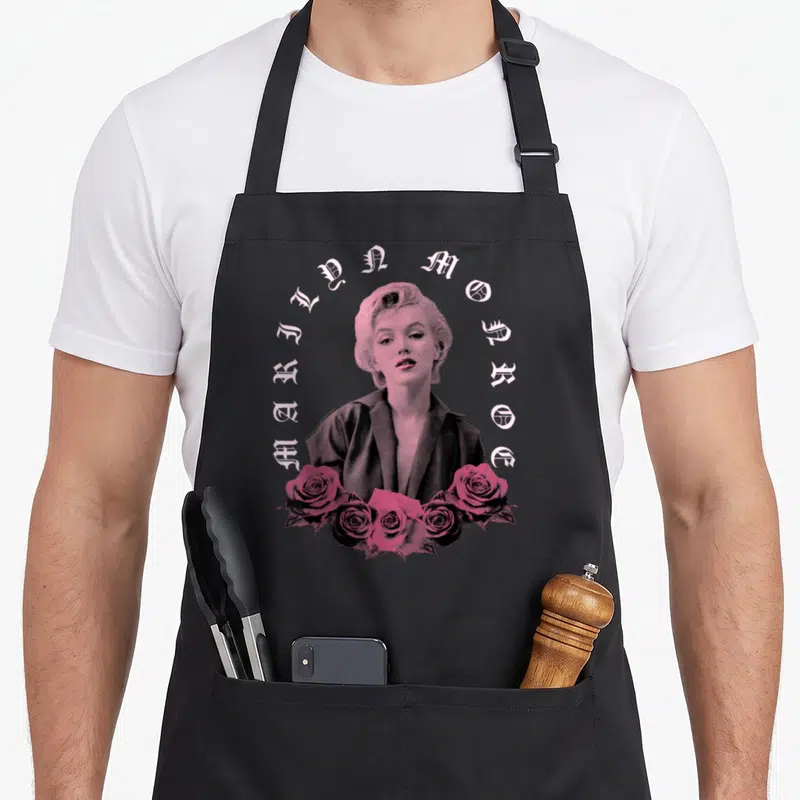 Marilyn english apron - Pink and grey Marilyn Monroe portrait with arched text and rose cluster — shown on a black cotton canvas apron against a white background