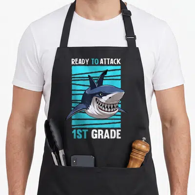 Ready school apron - Cartoon shark head on blue striped rectangle with ready to attack 1st grade text — shown on a black cotton canvas apron against a white background