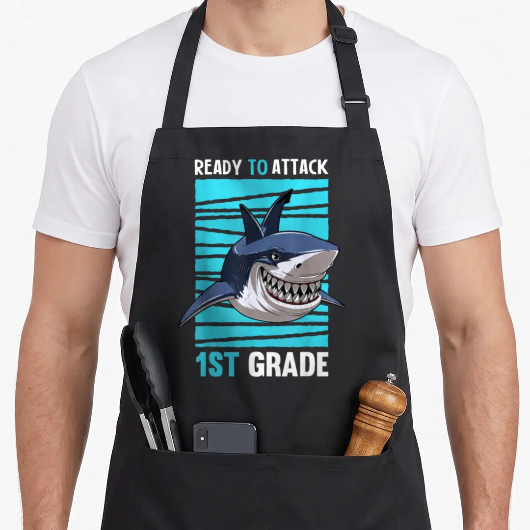 Ready school apron - Cartoon shark head on blue striped rectangle with ready to attack 1st grade text — shown on a black cotton canvas apron against a white background